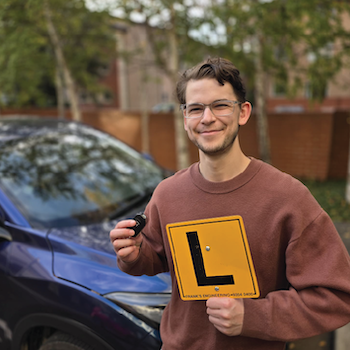 A learner driver smiling and holding an L-plate and car keys beside a vehicle.